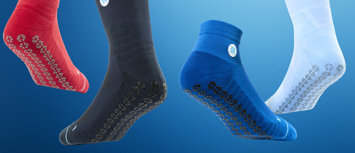 Four athletic grip socks floating against a blue gradient background: red, charcoal gray, bright blue, and white. Each sock shows textured non-slip dots on the sole and a small circular logo near the cuff.