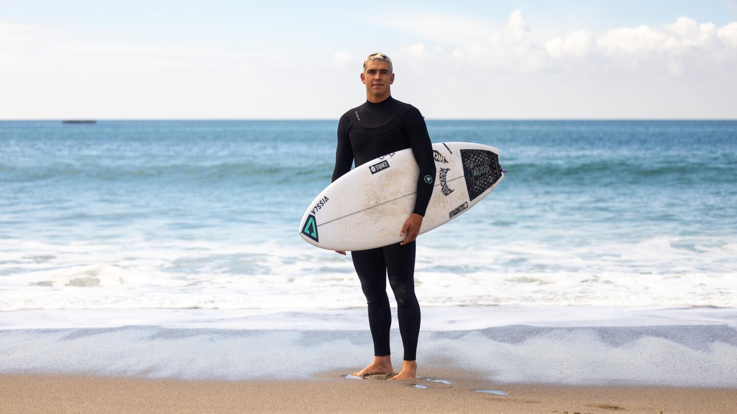 Photo of Cole Houshmand standing on the beach in front of the ocean wearing a black wet suit and holding a surfboard.