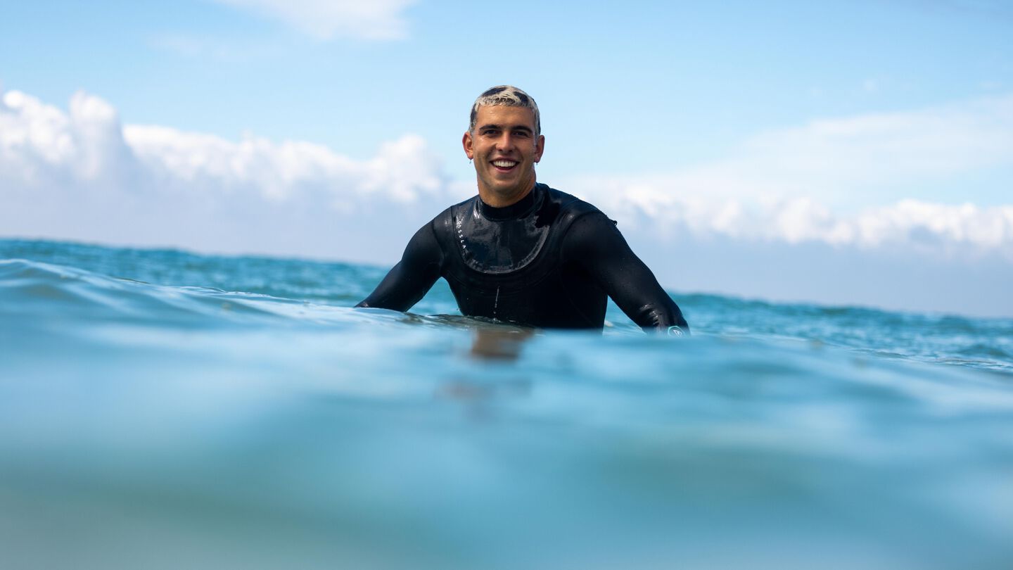 Photo of Cole Houshmand smiling while sitting on his surfboard in the ocean.